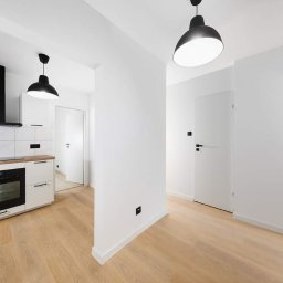 Adam Grygo - Jasne, nowocześnie wykończone wnętrze mieszkania z drewnianą podłogą, białą kuchnią z czarnymi akcentami i minimalistycznymi lampami wiszącymi.