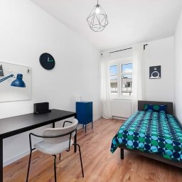 Adam Grygo - Pokój młodzieżowy z biurkiem, lampką kreślarską, łóżkiem jednoosobowym i geometryczną lampą sufitową, wykończony w bieli z akcentami koloru niebieskiego.