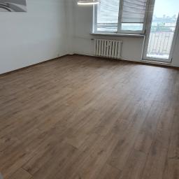 Adam Grygo - Pusty pokój z drewnianą podłogą, białymi ścianami, nowoczesnym, okrągłym, wiszącym oświetleniem i grzejnikiem pod oknem balkonowym z widokiem na miasto.