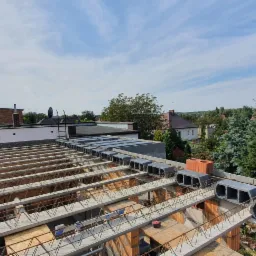 Wieniec stropowy z szalunkiem traconym wykonany z bloczków betonowych na budynku w stanie surowym otwartym, widok z góry na tle zabudowy jednorodzinnej i drzew.