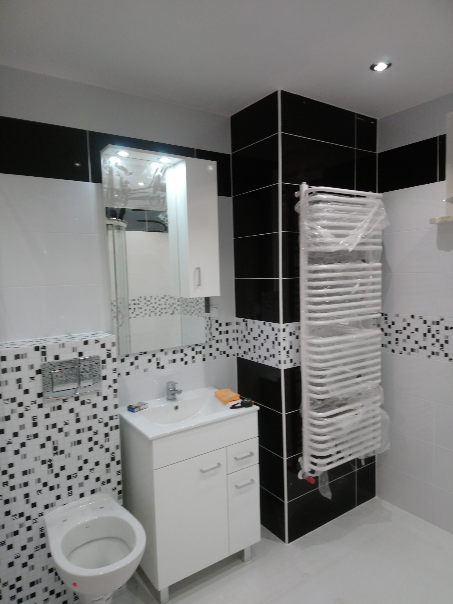 Nowoczesna łazienka z białą armaturą, czarno-białymi płytkami mozaikowymi i grzejnikiem drabinkowym w folii ochronnej, odzwierciedlającym światło w lustrze.