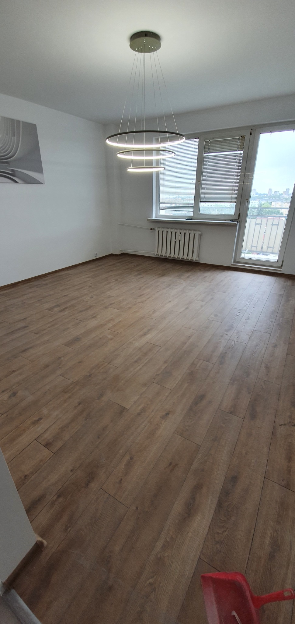 Pusty pokój z drewnianą podłogą, białymi ścianami, nowoczesnym, okrągłym, wiszącym oświetleniem i grzejnikiem pod oknem balkonowym z widokiem na miasto.