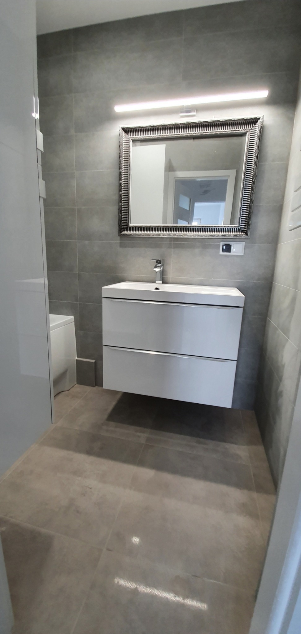 Nowoczesna łazienka z szarą glazurą, białą szafką pod umywalką z dwoma szufladami, lustrem w ozdobnej ramie i oświetleniem LED nad lustrem.