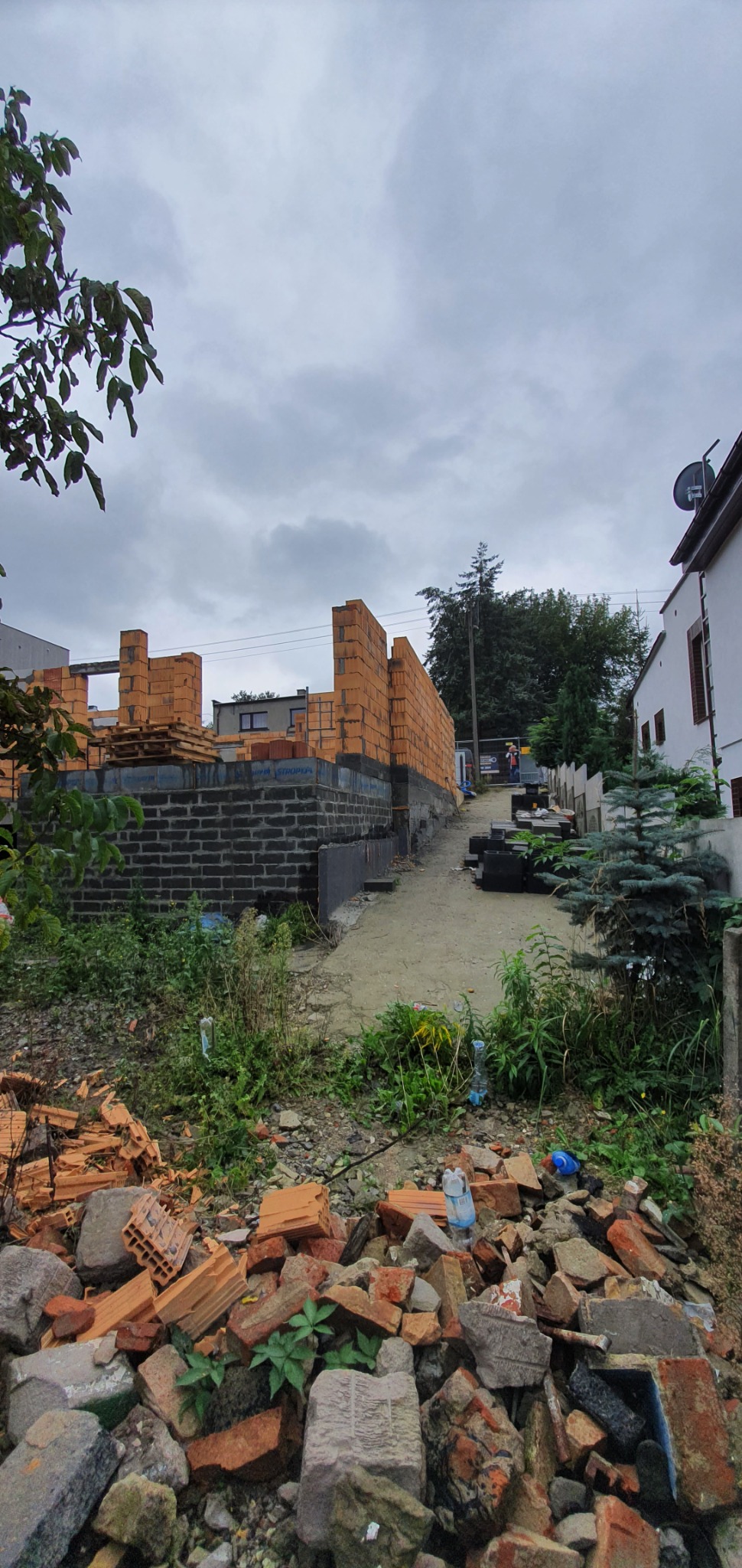 Wznoszone mury domu jednorodzinnego z bloczków ceramicznych, widok z dołu na plac budowy z gruzem i roślinnością.