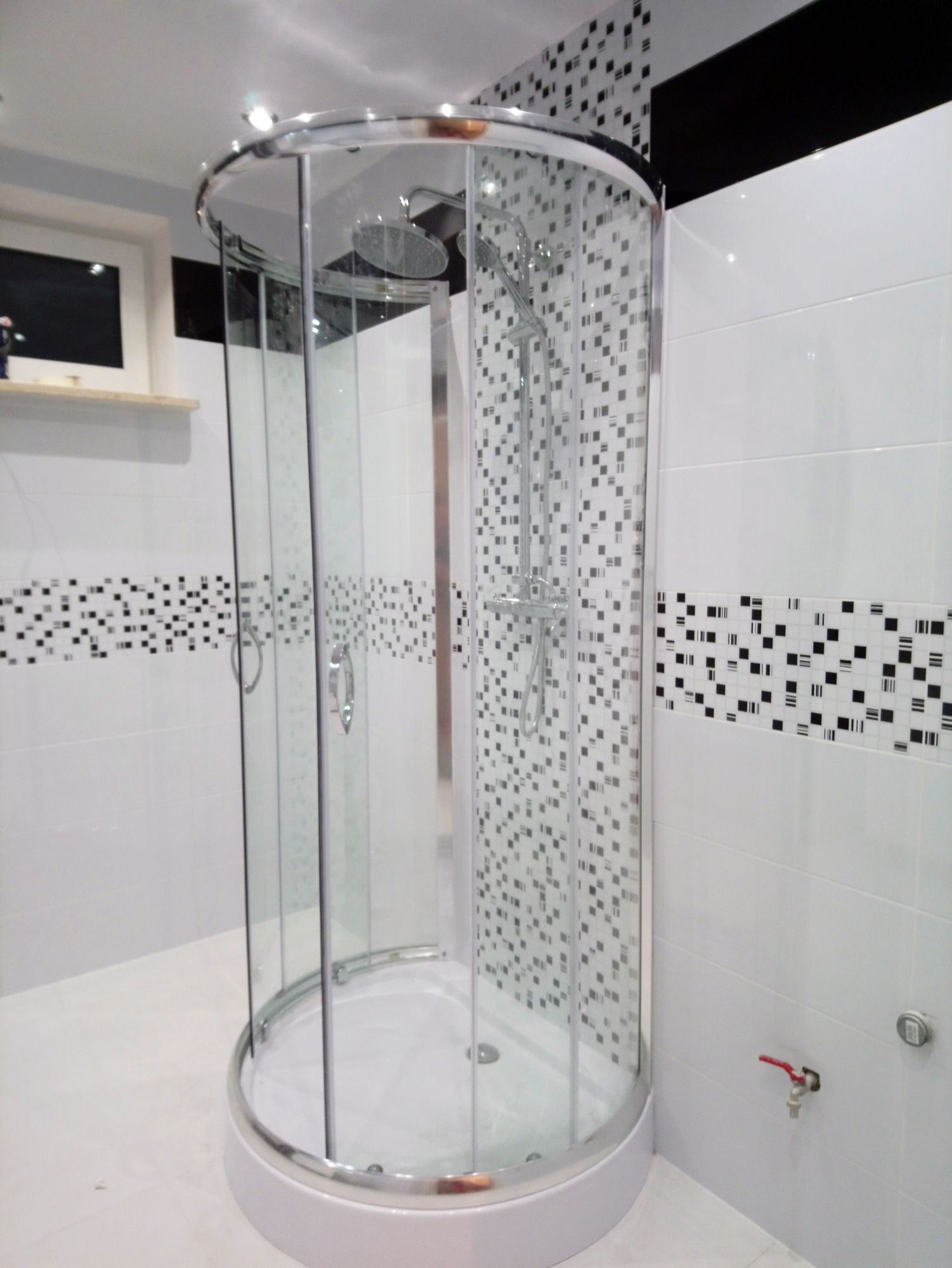 Zaokrąglona kabina prysznicowa z chromowanymi wykończeniami na tle białych płytek z dekoracyjnymi pasami czarno-białej mozaiki.