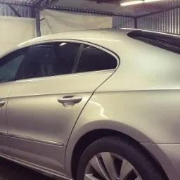 Srebrny samochód typu coupe z przyciemnionymi szybami bocznymi i tylną szybą, zaparkowany w warsztacie z widoczną konstrukcją sufitu.