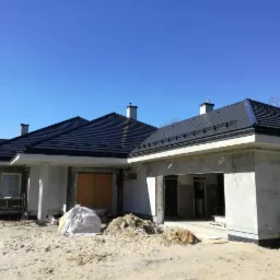 Stan surowy otwarty domu jednorodzinnego z ciemną dachówką, widoczny fundament i pracownik w zielonym kombinezonie przy wykopie, piasek na posesji.