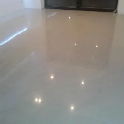 Świeżo ułożona, błyszcząca, beżowa podłoga z płytek ceramicznych, odbijająca światło od wbudowanego oświetlenia LED przy ścianie i światła z okna balkonowego.