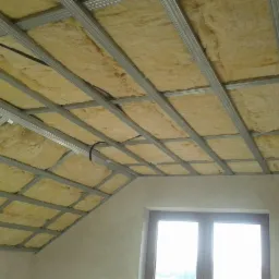 Skośny sufit poddasza z izolacją z wełny mineralnej umieszczoną w metalowej konstrukcji szkieletowej; widoczne okno w ścianie szczytowej.