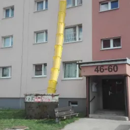 Żółty zsyp budowlany przymocowany do okna bloku mieszkalnego, prowadzący do kontenera na gruz na trawniku.