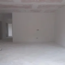 Wnętrze w trakcie prac wykończeniowych: surowy sufit z widocznymi łączeniami płyt, ściany po nałożeniu pierwszej warstwy, gładka, jasna podłoga z niewielkim wiadrem na pierwszym planie, widoczne...