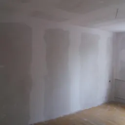 Pokój w trakcie prac wykończeniowych; ściany z widocznymi łączeniami płyt, przygotowane do dalszej obróbki; drewniana podłoga; widoczne fragmenty instalacji grzewczej.