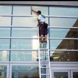 Pracownik na drabinie myje okna budynku biurowego za pomocą ściągaczki, widoczne odbicie budynku w szybach.