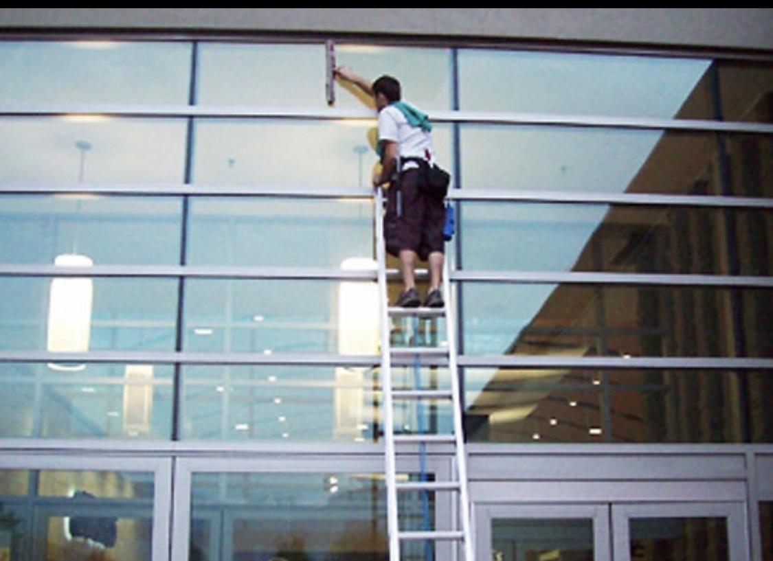 Pracownik na drabinie myje okna budynku biurowego za pomocą ściągaczki, widoczne odbicie budynku w szybach.
