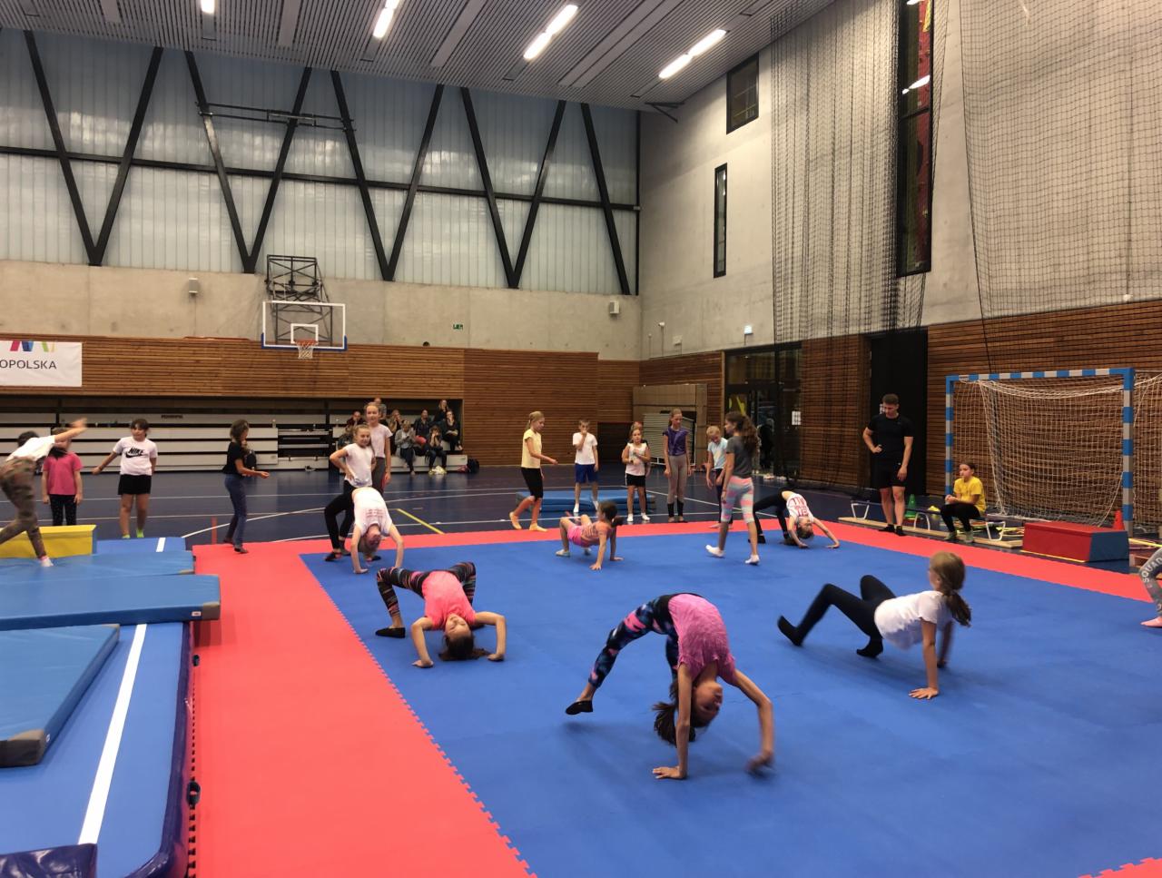 Grupowe zajęcia gimnastyczne dla dzieci w hali sportowej, z dziewczynkami wykonującymi mostki i inne ćwiczenia na niebiesko-czerwonej macie pod nadzorem instruktora.