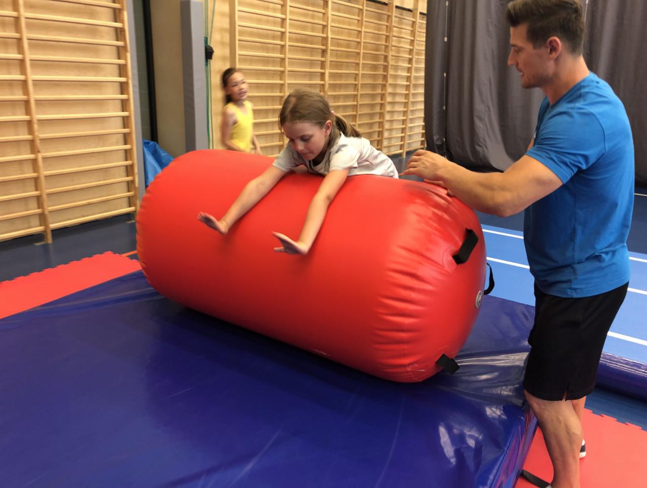 Dziewczynka ćwiczy na czerwonym walcu gimnastycznym, a instruktor pomaga jej w sali gimnastycznej z drewnianą ścianą i niebiesko-czerwoną podłogą.