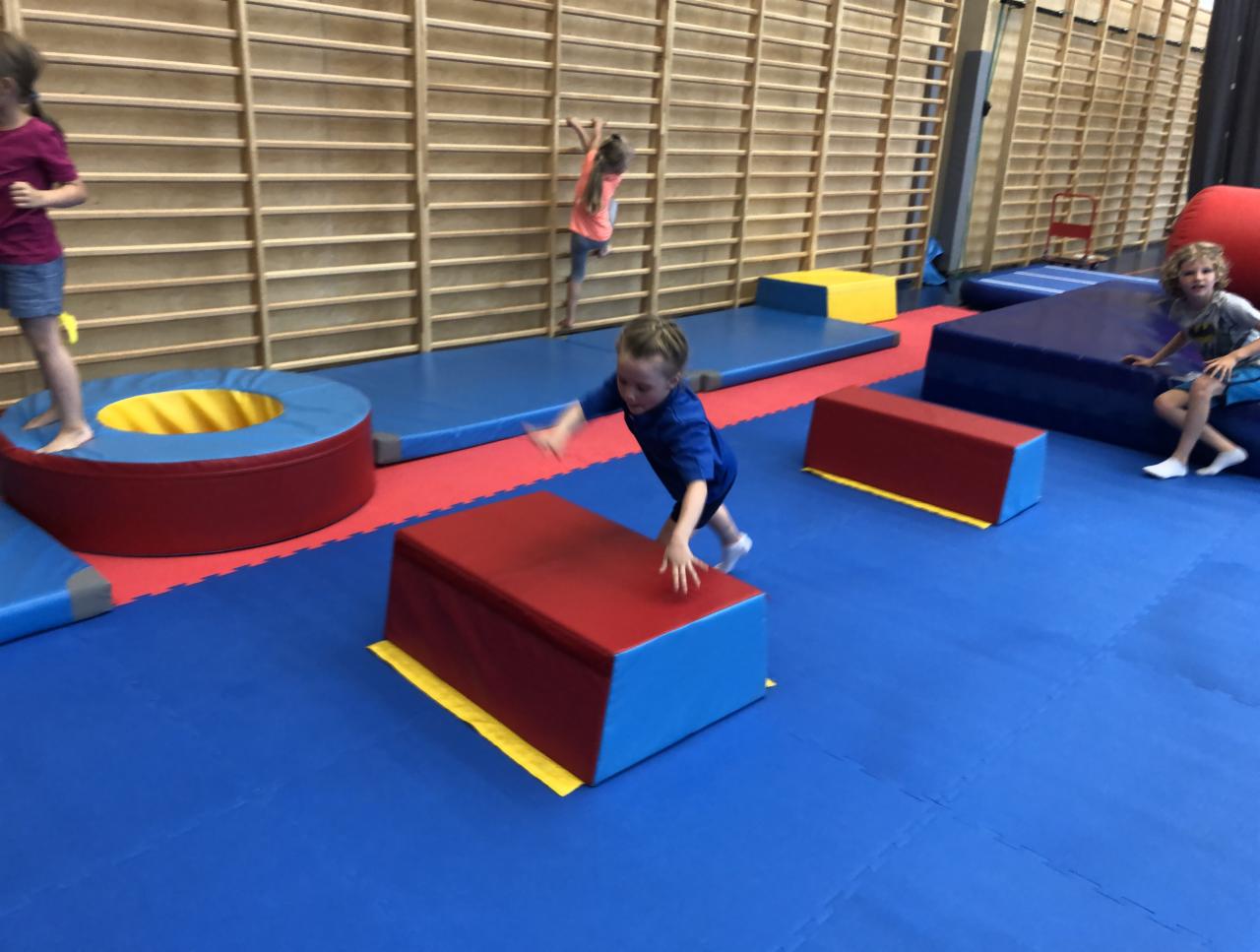 Chłopiec w niebieskiej koszulce skacze przez czerwony materac gimnastyczny na tle ściany z drabinkami i innych dzieci ćwiczących w sali gimnastycznej.