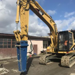 Żółta koparka gąsienicowa CAT 318B z niebieskim młotem hydraulicznym na tle budynku z brązowymi wrotami garażowymi i częściowo pochmurnego nieba.