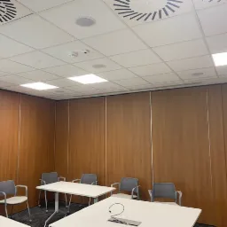 Sala konferencyjna z drewnianymi panelami ściennymi, białym sufitem kasetonowym, okrągłymi kratkami wentylacyjnymi i kwadratowymi lampami, widoczne białe stoły i szare krzesła.