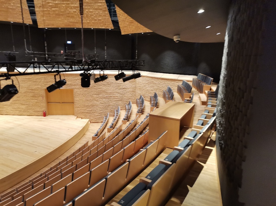 Wnętrze sali koncertowej z rzędami foteli i oświetleniem technicznym zawieszonym pod sufitem, widok z góry na scenę i widownię z drewnianymi elementami wykończeniowymi.