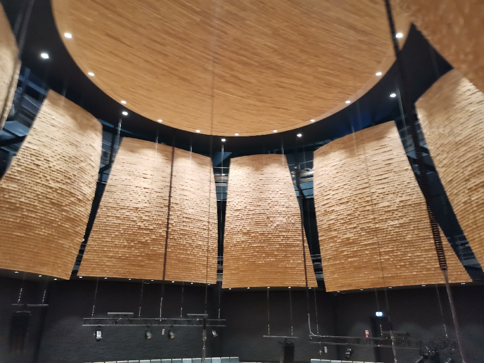 Widok z dołu na drewniane panele akustyczne zamontowane na suficie i ścianach sali koncertowej, oświetlone punktowymi lampami.