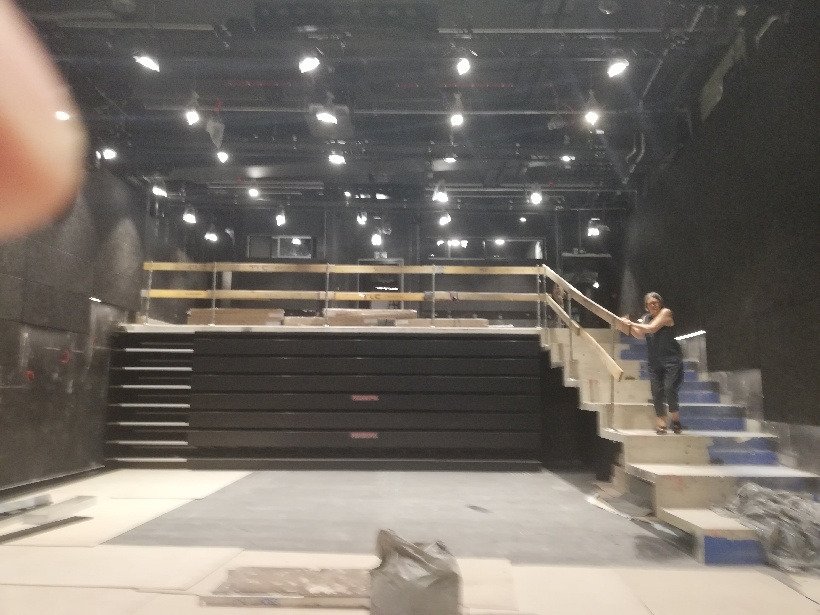 Widok sali teatralnej w trakcie prac instalacyjnych oświetlenia, widoczne reflektory podwieszone pod sufitem i drabina z osobą na schodach.