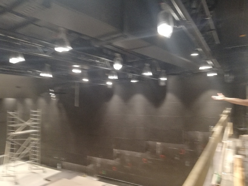 Wysokie pomieszczenie z zamontowanym oświetleniem sufitowym, widoczny fragment rusztowania i ręka wskazująca na instalację. Oświetlenie punktowe skierowane na ścianę.