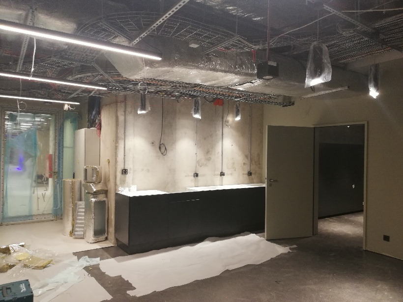 Wnętrze w trakcie prac wykończeniowych: widoczne kable pod sufitem, ściany w surowym betonie, czarna zabudowa meblowa, podłoga zabezpieczona folią i otwarte drzwi do kolejnego pomieszczenia.