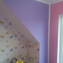 Pokój dziecięcy po malowaniu: ściana w kolorze liliowym sąsiaduje z różową, a skośna ściana poddasza pokryta jest tapetą w dziecięce wzory. Żółty odkurzacz stoi w rogu, a fragment drabiny wskazuje...