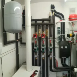 Złożona instalacja grzewcza z licznymi rurami, zaworami i pompami, w pomieszczeniu technicznym o białych ścianach, widoczny manometr i zbiornik wyrównawczy.