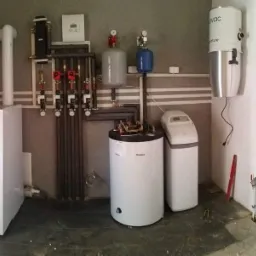 Kompletna kotłownia z kotłem gazowym, zasobnikiem ciepłej wody, zmiękczaczem wody i rekuperatorem centralnym na ścianie, widoczne rury i armatura.