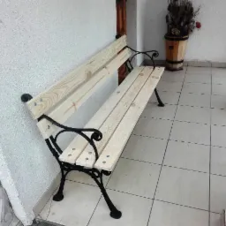 Nowa, surowa ławka ogrodowa z jasnego drewna i czarnymi, metalowymi nogami, ustawiona na wyłożonym płytkami tarasie przy białej ścianie budynku. W tle widoczna donica z roślinami.