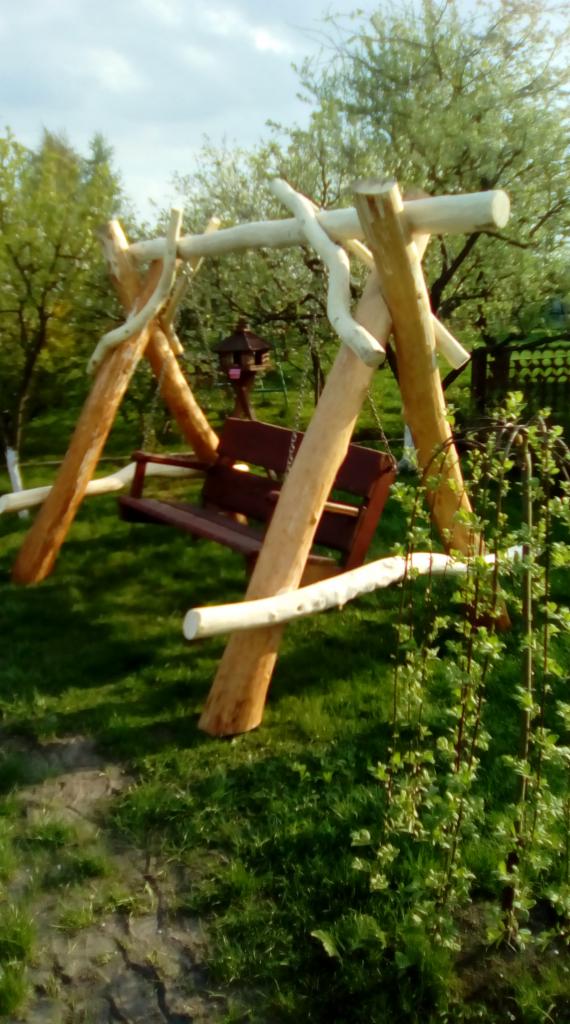 Drewniana huśtawka ogrodowa z siedziskiem w kolorze brązowym, wykonana z surowych bali, w ogrodzie w Kozłowie. W tle widoczne drzewa i karmnik dla ptaków.