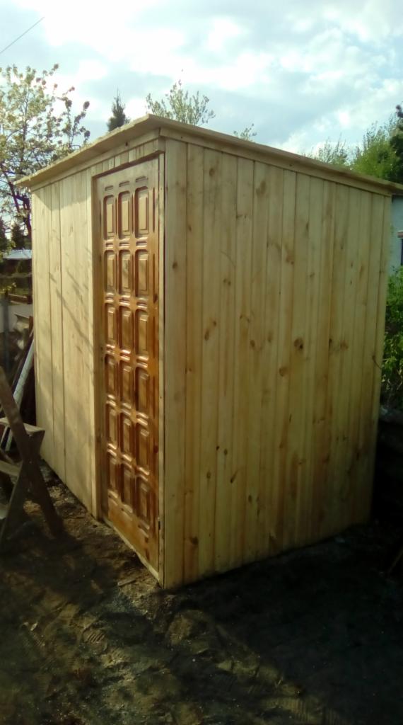 Prosta, drewniana konstrukcja z ciemniejszymi, rzeźbionymi drzwiami, prawdopodobnie budka lub mały schowek, umieszczona na ziemi z widocznymi śladami prac budowlanych.