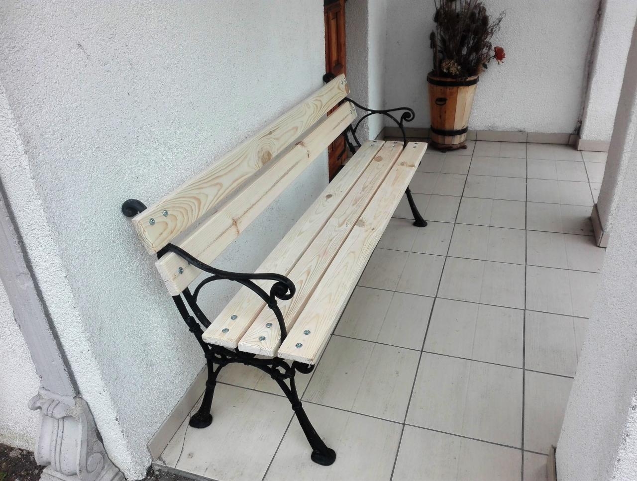 Nowa, surowa ławka ogrodowa z jasnego drewna i czarnymi, metalowymi nogami, ustawiona na wyłożonym płytkami tarasie przy białej ścianie budynku. W tle widoczna donica z roślinami.