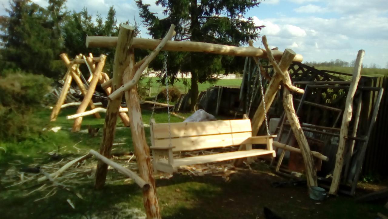 Niedokończona drewniana huśtawka ogrodowa z bali, widoczne łańcuchy, w tle druga huśtawka w trakcie budowy i elementy drewniane do konstrukcji.