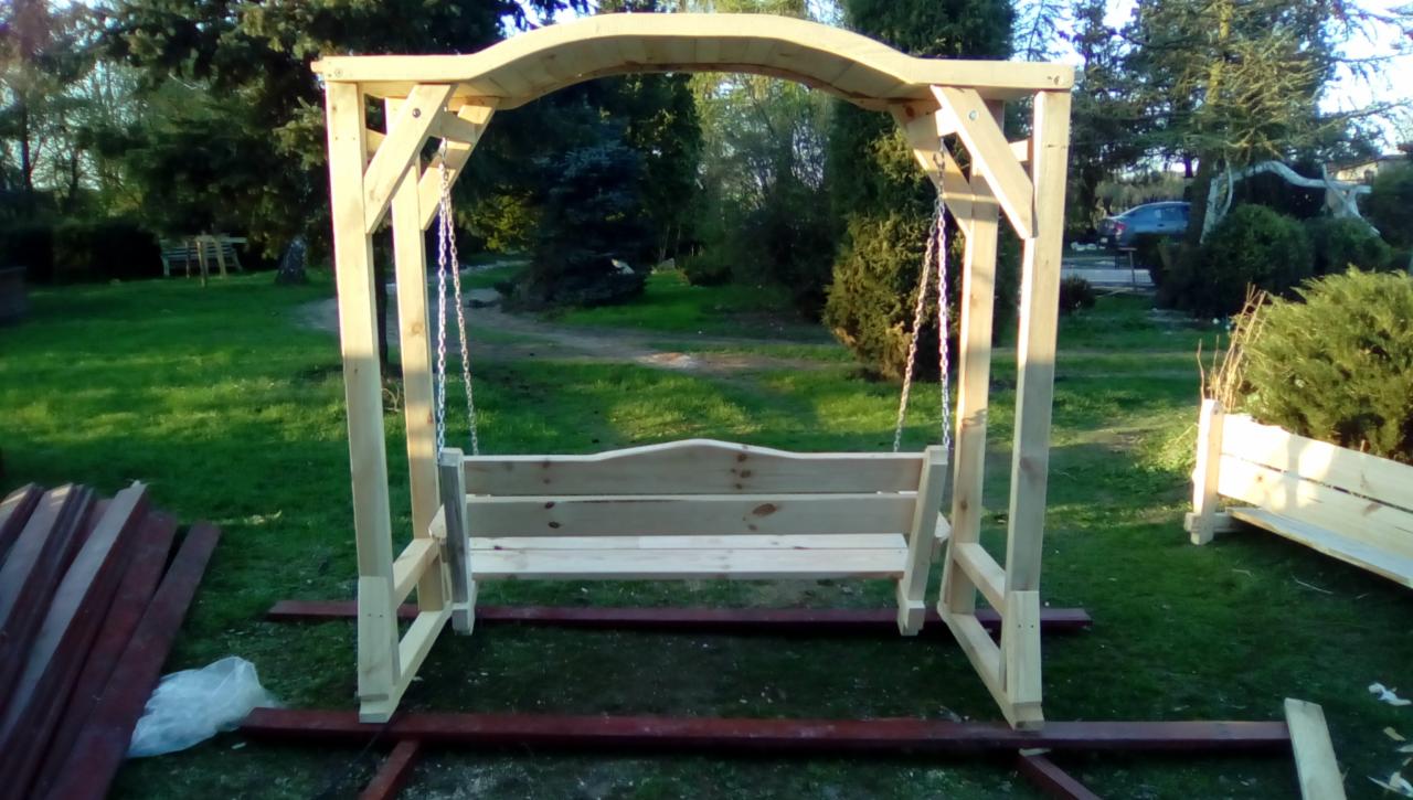 Niedokończona drewniana huśtawka ogrodowa z zadaszeniem łukowym na łańcuchach, ustawiona na trawie w ogrodzie, widoczne deski i elementy konstrukcyjne w pobliżu.
