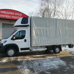 Biały bus z plandeką na tle budynku Okręgowej Stacji Kontroli Pojazdów i resztek śniegu na chodniku.