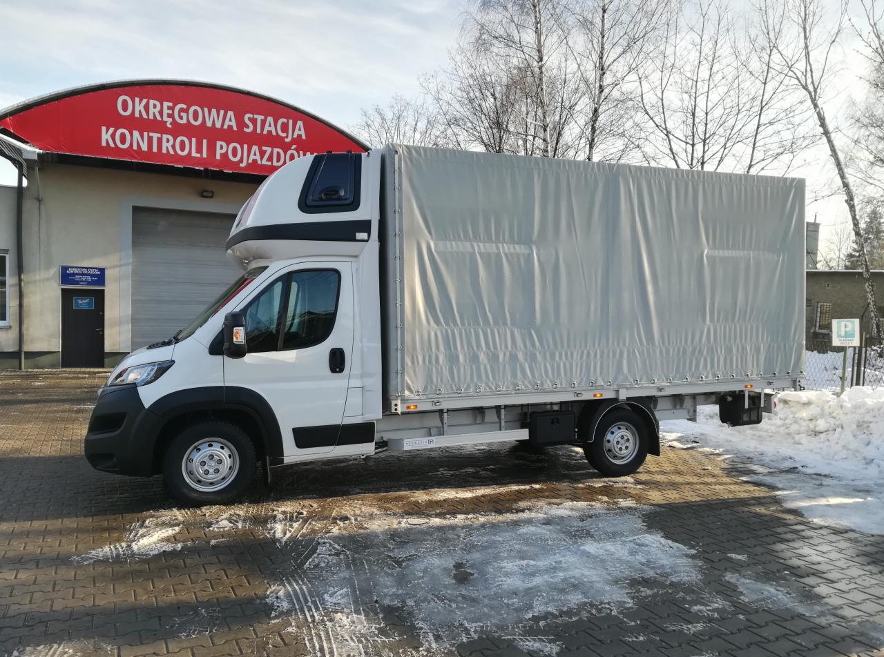 Biały bus z plandeką na tle budynku Okręgowej Stacji Kontroli Pojazdów i resztek śniegu na chodniku.