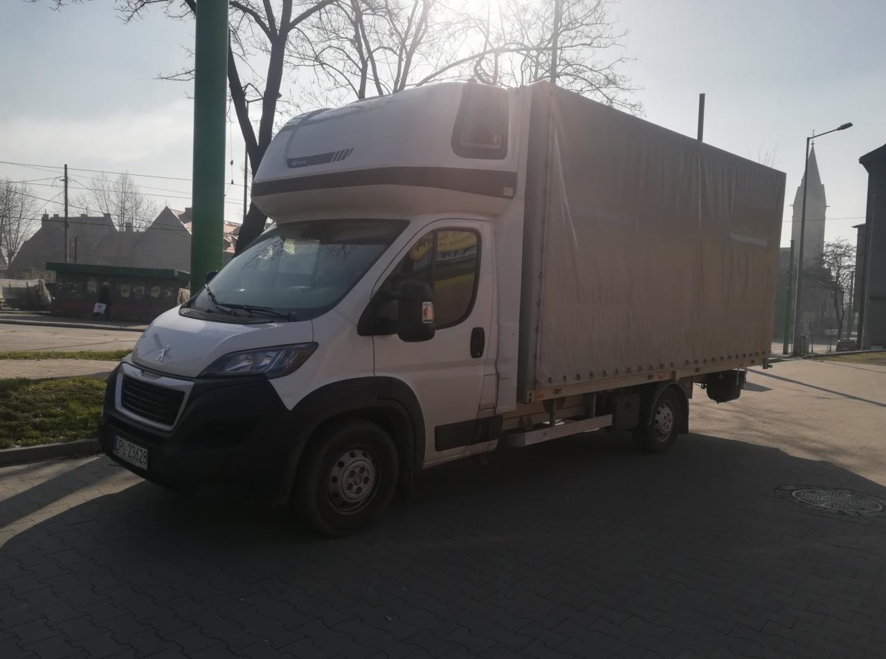 Biały bus dostawczy z plandeką zaparkowany na brukowanej ulicy w słoneczny dzień, z widocznym kościołem w tle.