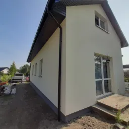 Nowo otynkowana elewacja domu jednorodzinnego z ciemnymi rynnami, widok z boku na jasnym tle nieba, z materiałami budowlanymi i samochodem dostawczym w tle.