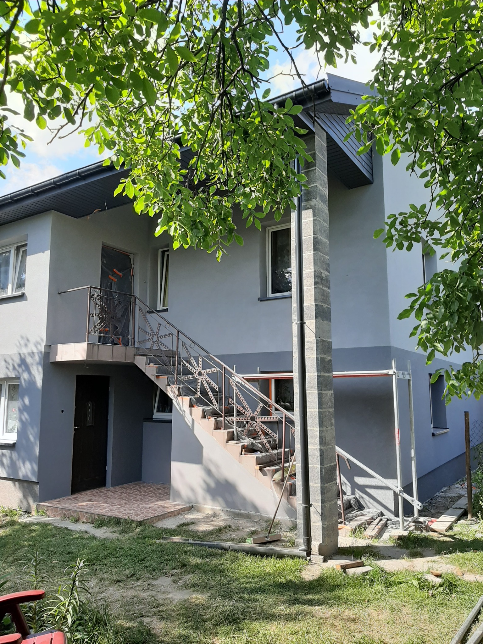 Dwu kondygnacyjny dom z szarą elewacją, widoczne schody zewnętrzne z metalową balustradą prowadzące do drzwi na piętrze, okna zabezpieczone folią, fragment rusztowania, w tle zieleń drzew