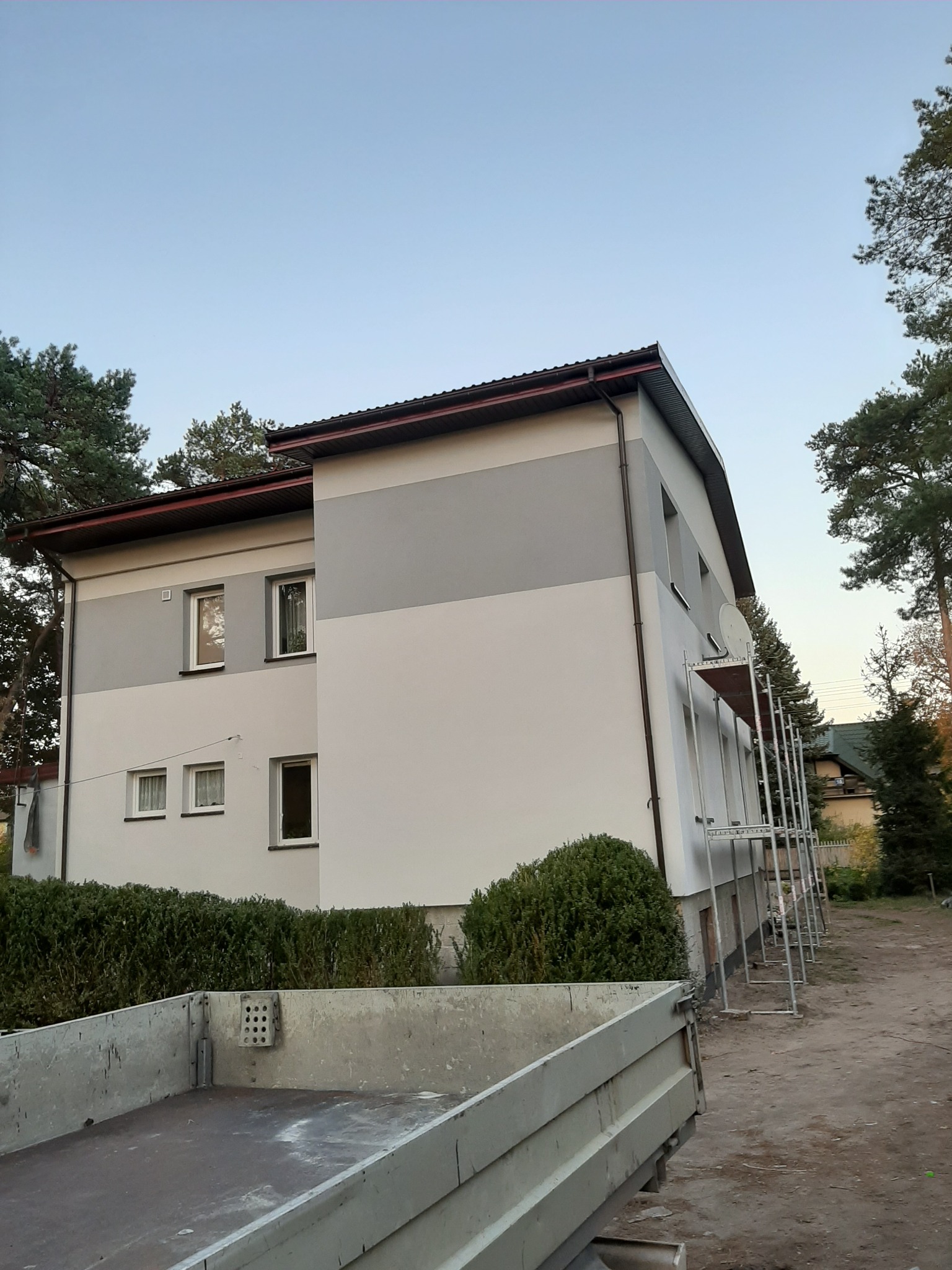 Dwukondygnacyjny dom po renowacji elewacji z poziomym podziałem kolorystycznym (szary/biały), z widocznymi rusztowaniami i skrzynią ładunkową pojazdu na pierwszym planie.