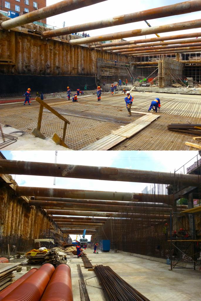 Ekipa budowlana w pomarańczowych kaskach i niebieskich uniformach pracuje nad zbrojeniem fundamentów w głębokim wykopie zabezpieczonym stalowymi rurami i ściankami berlińskimi.