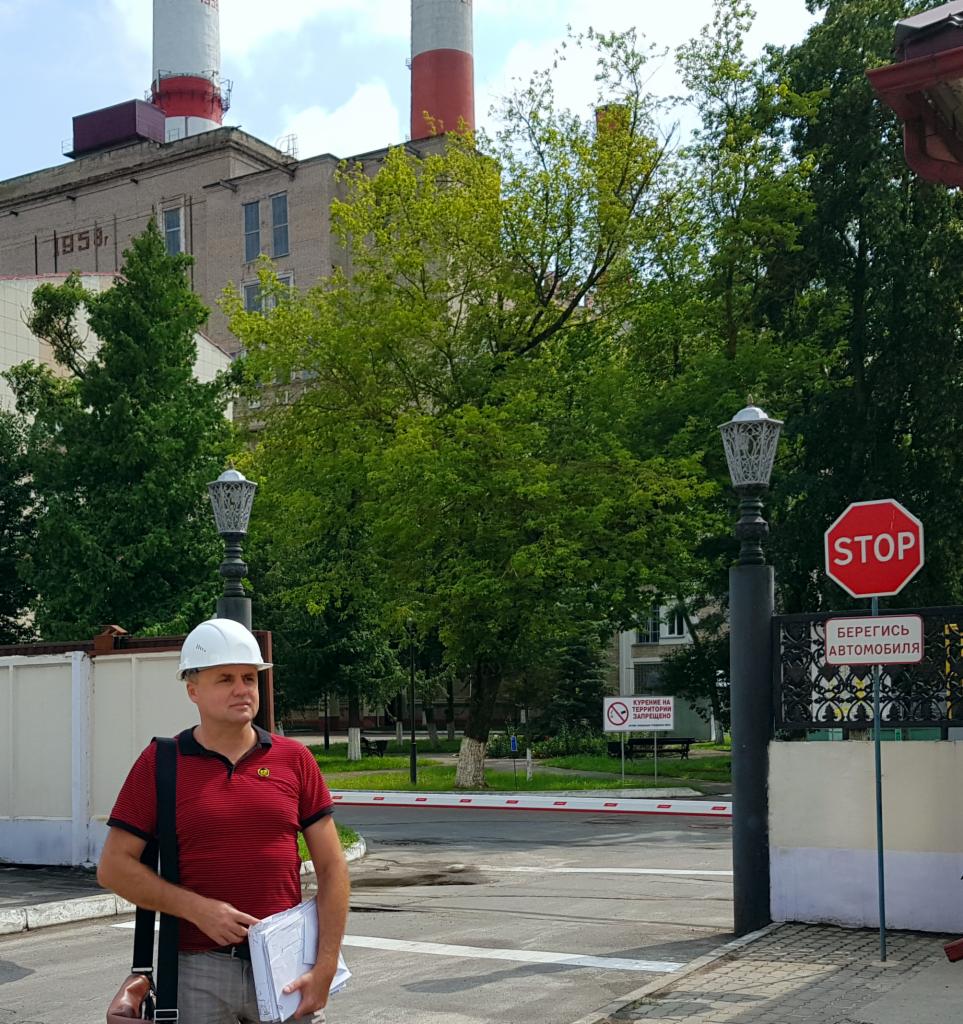 Mężczyzna w kasku, trzymający dokumenty, stoi przed wjazdem na teren przemysłowy z widoczną elektrownią w tle, znak STOP oraz barierka.