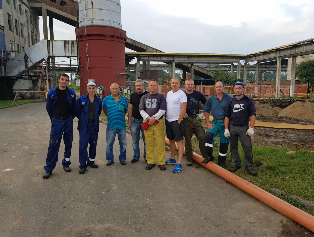 Grupa dziewięciu pracowników w różnej odzieży roboczej pozuje na tle budowy z rurą u dołu kadru, widoczny budynek przemysłowy w tle.