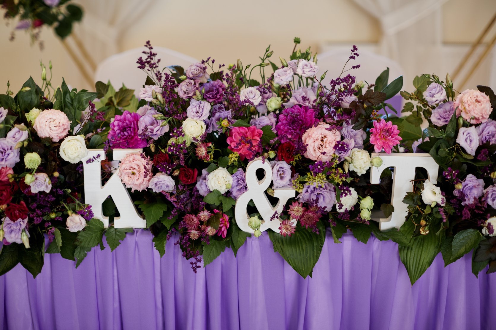 Dekoracja stołu weselnego z kwiatów w odcieniach fioletu i różu, z białymi literami K i T oraz znakiem '&', na liliowym obrusie.