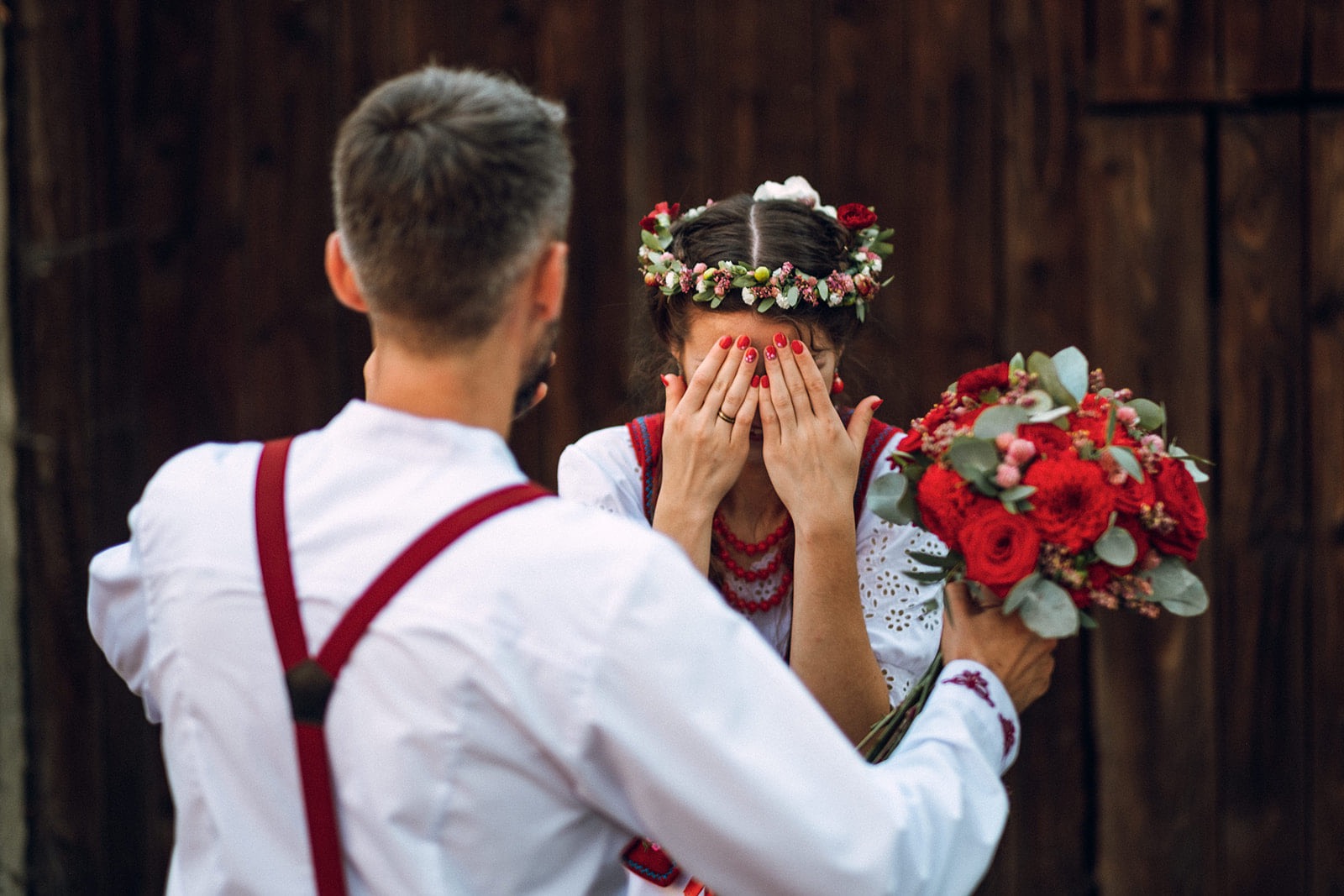 Mężczyzna wręcza bukiet czerwonych róż kobiecie w ludowym stroju z wiankiem na głowie, która zasłania twarz dłońmi, na tle drewnianej ściany.