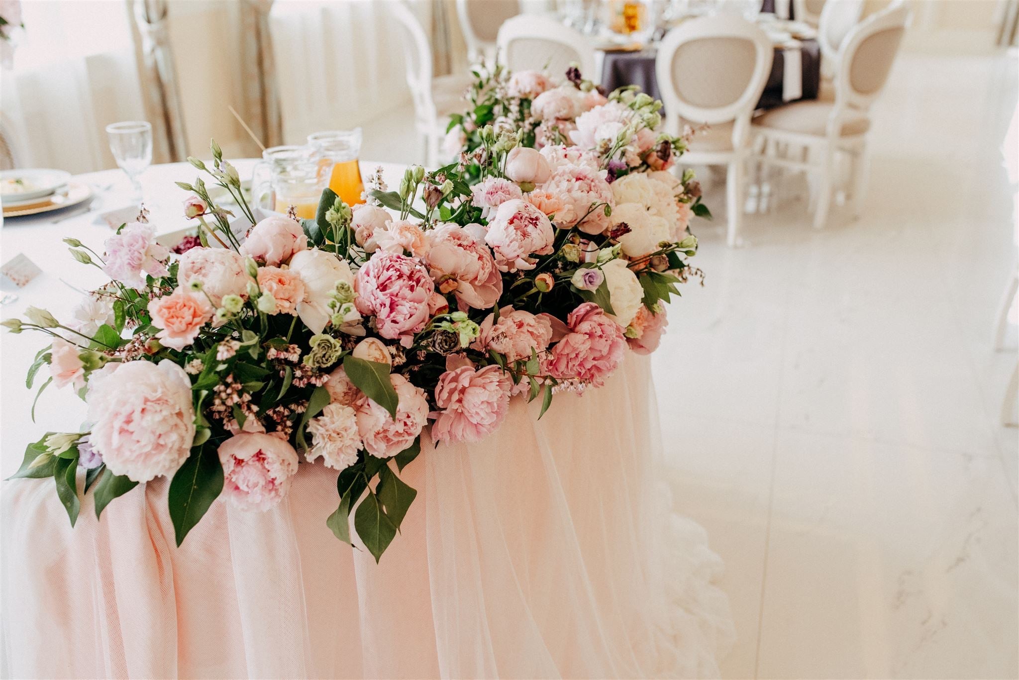 Dekoracja stołu z różowych i białych piwonii, goździków i zielonych liści, z widocznymi szklankami i dzbankiem z sokiem w tle, w jasnym, eleganckim wnętrzu.