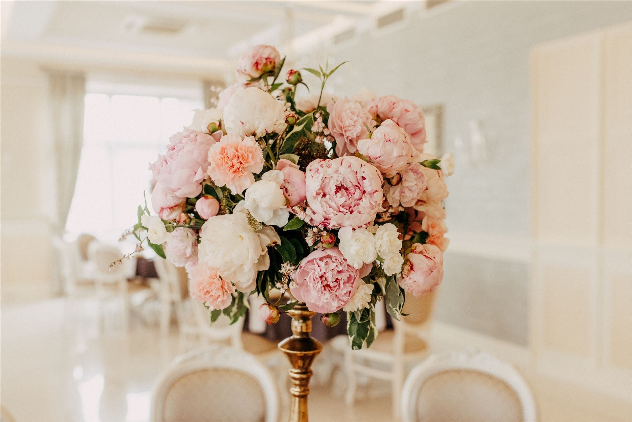 Dekoracyjny bukiet z piwonii, goździków i drobnych białych kwiatów w odcieniach bieli i różu, umieszczony w złotym, metalowym wazonie na stole w jasnym wnętrzu.